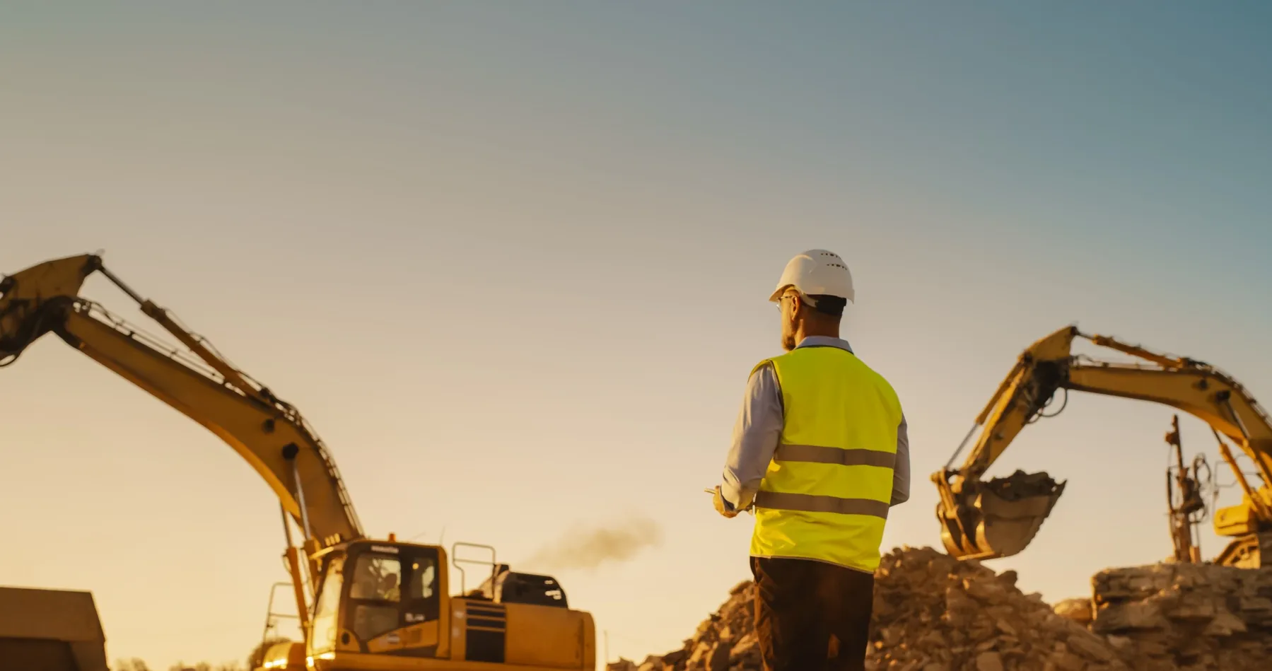 Construction worker observing excavators at sunset.