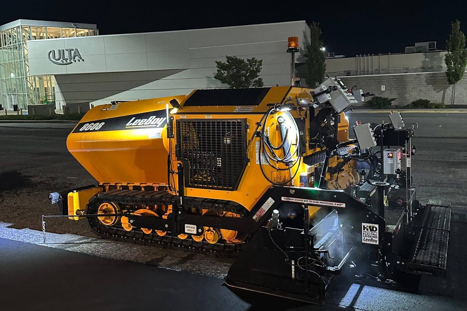 Asphalt paving machine, LeeBoy 9000 model, illuminated at night in front of an Ulta Beauty store, showcasing industrial paving equipment for commercial sites.