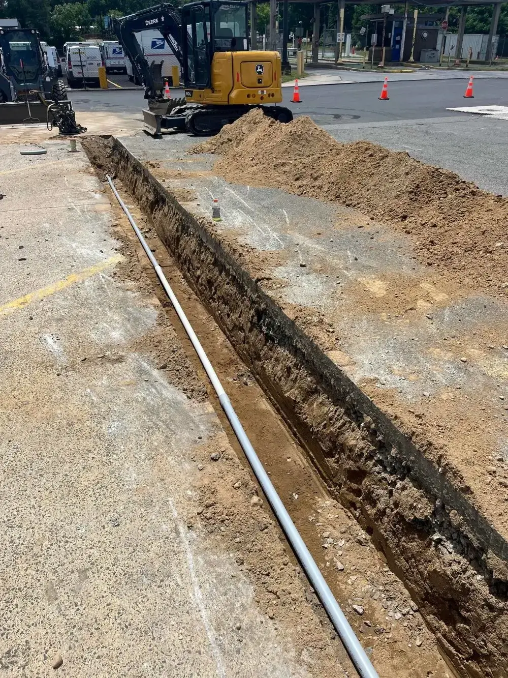 Excavated trench for utility installation with a yellow compact excavator and sand piles, showcasing groundwork for asphalt paving and concrete restoration projects.