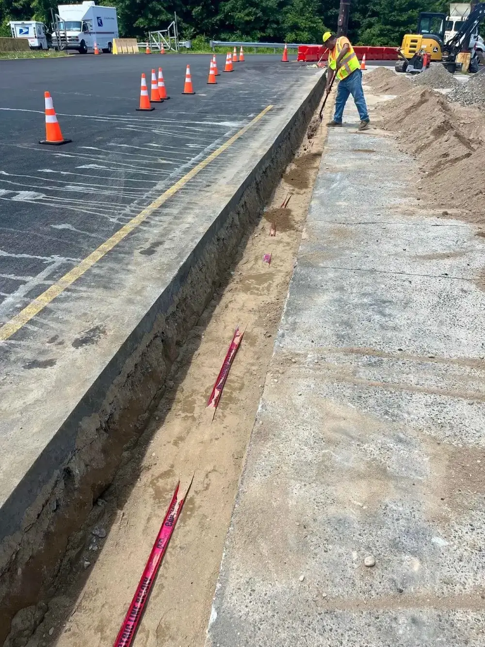 Construction worker excavating trench for asphalt paving project, marked with safety cones and measuring tape, showcasing Conte Paving & Construction's expertise in site work and concrete restoration.